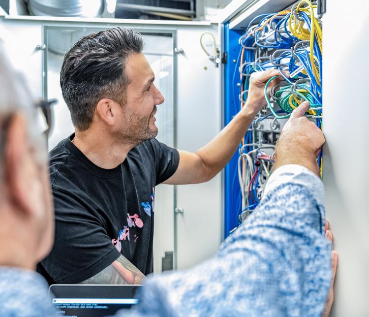 Een man in een zwarte T-shirt werkt aan een netwerkserverkast en is bezig met het organiseren van kabels, terwijl een collega op de voorgrond toekijkt en ondersteunt.