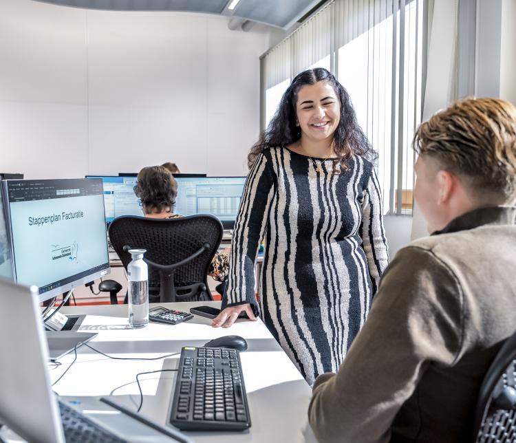Een vrouw in een gestreepte jurk staat lachend in gesprek met een collega in een moderne kantoorruimte met werkplekken en computers. Op een van de schermen is een presentatie te zien met de titel 'Stappenplan Facturatie'