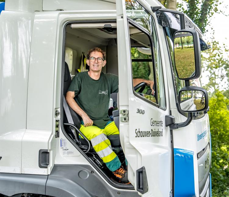 Een man in werkkleding zit glimlachend achter het stuur in een vrachtwagen van de gemeente Schouwen-Duiveland, met de deur open en een groene, bosrijke omgeving op de achtergrond.
