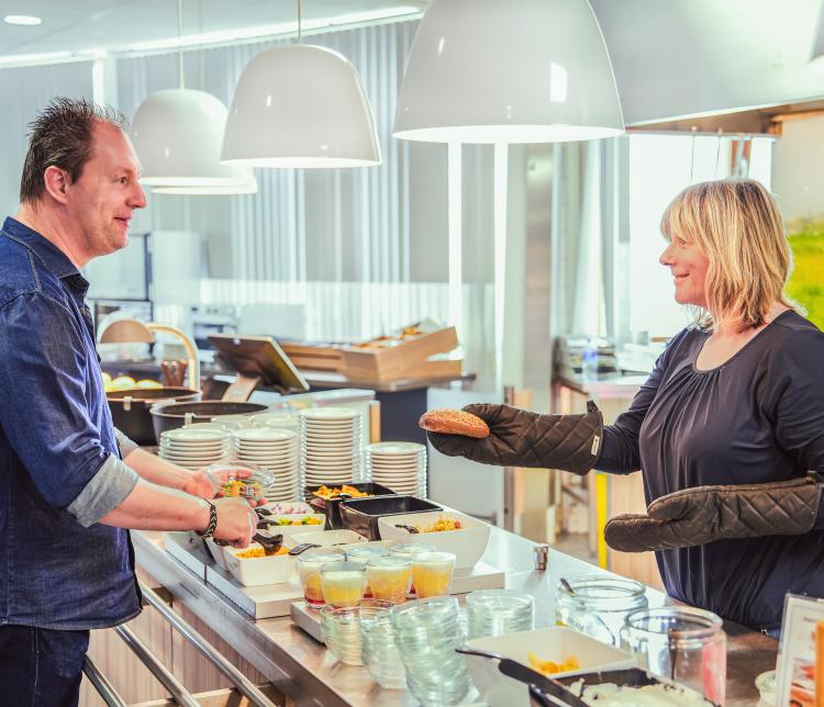 Een medewerker van het bedrijfsrestaurant reikt een vers broodje aan een klant, terwijl deze een salade opschept. De moderne eetruimte heeft een uitnodigende en goed georganiseerde uitstraling.