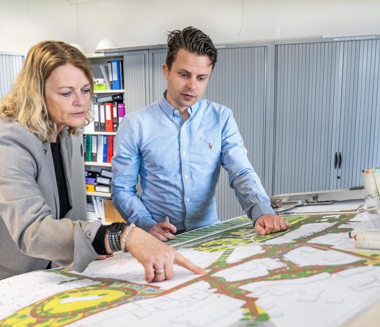 Een man en een vrouw staan gebogen over een grote plattegrond op een bureau in een kantoorruimte. De vrouw in een grijs jasje wijst naar een specifiek deel van de kaart, terwijl de man in een lichtblauw overhemd aandachtig meekijkt en een marker vasthoudt. Op de tafel liggen diverse stiften en tekengerei. Op de achtergrond zijn archiefkasten en kleurrijke ordners te zien.