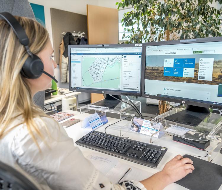 Medewerker van de gemeente Schouwen-Duiveland zit met een headset op achter een bureau en werkt aan twee computerschermen. Op het linkerscherm staat een kaartweergave, op het rechterscherm de website van de gemeente. Op het bureau liggen notities, een toetsenbord en een kalender.