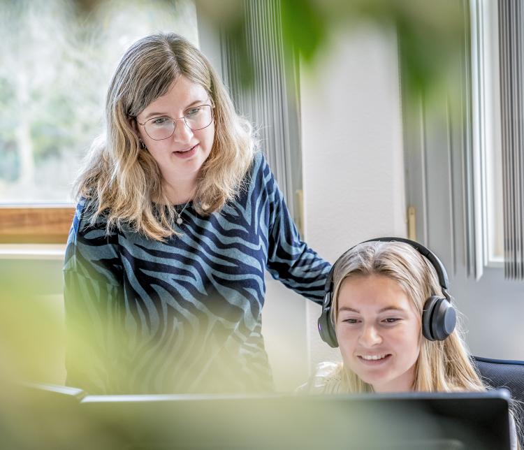 Twee vrouwelijke collega’s bij de gemeente Schouwen-Duiveland werken samen achter een computerscherm. De jongere vrouw draagt een headset en zit, terwijl de andere vrouw naast haar staat en meekijkt. Ze bevinden zich in een lichte kantoorruimte met grote ramen en planten op de voorgrond.