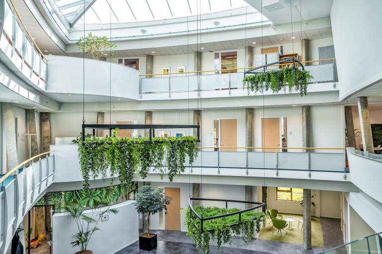 Een modern atrium van een kantoorruimte met meerdere verdiepingen, voorzien van glazen balustrades en hangende groene plantenbakken. Het dak is gemaakt van glas, wat veel natuurlijk licht binnenlaat. Op de begane grond staan enkele groene stoelen en een ronde tafel, terwijl grote planten en kunstzinnige muurschilderingen het interieur sieren. Betonpilaren en houten accenten geven de ruimte een moderne uitstraling.