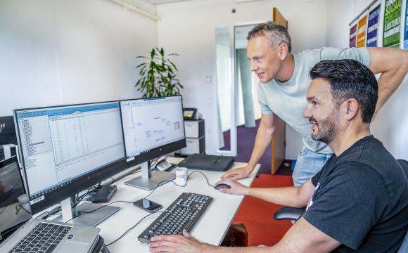 Twee mannen werken samen in een kantooromgeving. Eén man zit achter een bureau met meerdere computerschermen en toetsenbord, terwijl de andere man naast hem staat en meedenkt. Ze bekijken gegevens en diagrammen op de schermen.
