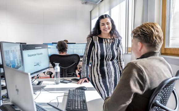 Een vrouw in een gestreepte jurk staat lachend in gesprek met een collega in een moderne kantoorruimte met werkplekken en computers. Op een van de schermen is een presentatie te zien met de titel 'Stappenplan Facturatie'