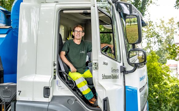 Een man in werkkleding zit glimlachend achter het stuur in een vrachtwagen van de gemeente Schouwen-Duiveland, met de deur open en een groene, bosrijke omgeving op de achtergrond.
