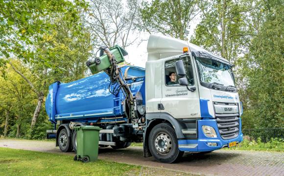 Een vuilniswagen van de gemeente Schouwen-Duiveland is bezig met het legen van een groene afvalcontainer met een automatische grijparm, in een groene, bosrijke omgeving. De chauffeur zit glimlachend achter het stuur.