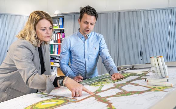 Een man en een vrouw staan gebogen over een grote plattegrond op een bureau in een kantoorruimte. De vrouw in een grijs jasje wijst naar een specifiek deel van de kaart, terwijl de man in een lichtblauw overhemd aandachtig meekijkt en een marker vasthoudt. Op de tafel liggen diverse stiften en tekengerei. Op de achtergrond zijn archiefkasten en kleurrijke ordners te zien.