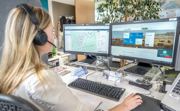 Medewerker van de gemeente Schouwen-Duiveland zit met een headset op achter een bureau en werkt aan twee computerschermen. Op het linkerscherm staat een kaartweergave, op het rechterscherm de website van de gemeente. Op het bureau liggen notities, een toetsenbord en een kalender.