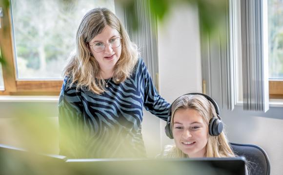 Twee vrouwelijke collega’s bij de gemeente Schouwen-Duiveland werken samen achter een computerscherm. De jongere vrouw draagt een headset en zit, terwijl de andere vrouw naast haar staat en meekijkt. Ze bevinden zich in een lichte kantoorruimte met grote ramen en planten op de voorgrond.